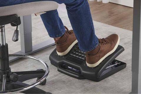 Pés com sapato marrom descansando em cima de uma base de apoio que está em baixo da mesa